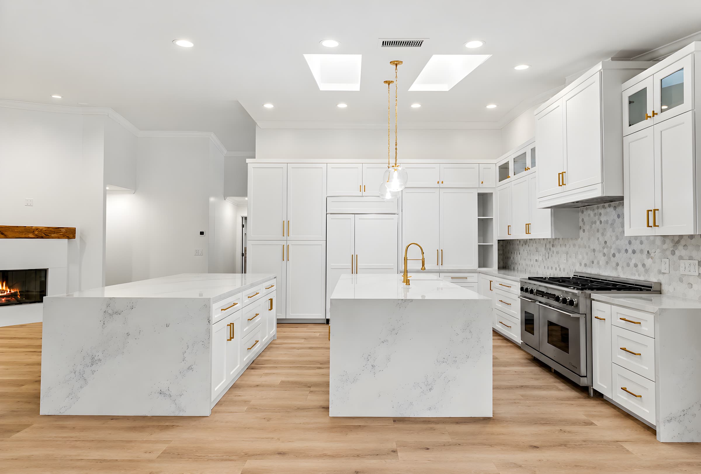 Completed kitchen with twin marble waterfall islands, brass-handled white shaker cabinets, mosaic backsplash, and a six-burner range under a custom hood.